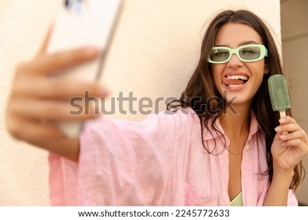 Happy young caucasian woman in sunglasses takes selfie on smartphone holds ice cream. Brunette wears pink shirt and green sundress. Use, device concept.