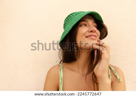 Close-up of nice young caucasian woman lookis away smiles stands near peach color wall. Model with brunette hair wears green panama hat and sundress. Concept of great mood.
