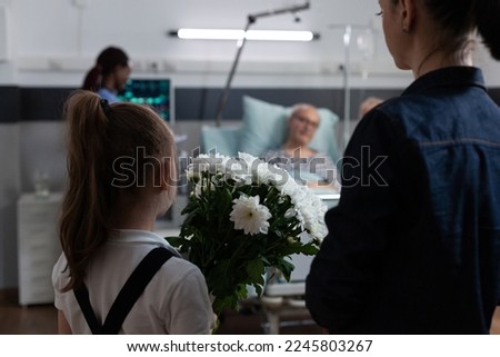Woman, daughter arriving with flowers at sick elderly man hospital room. African american doctor checking bedridden old patient vital signs. Girl holding floral arrangement for hospitalized grandpa.