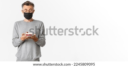 Concept of covid-19, quarantine and holidays. Image of dreamy middle-aged man in medical mask and glasses, looking amazed at birthday cake, making a wish, standing over white background.