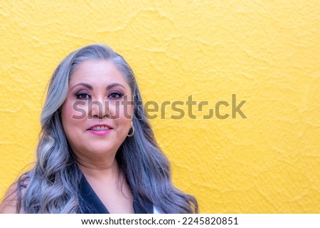 Frontal portrait of senior adult woman looking at camera over yellow background, grey-black hair, warm color makeup, lips with pink lipstick, formal wear, black and white blouse