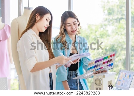 Millennial Asian young professional female dressmaker designer seamstress with measuring tape and colleague partner help checking color pattern with drawing sketching in tailor studio workshop office.