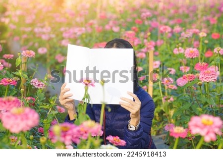 Teenage girl covering her face with blank book or magazine with blooming pink zinnia flowers field, people with in landscape.