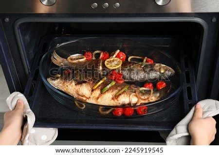 Woman taking out baking tray with sea bass fish and garnish from oven, closeup Royalty-Free Stock Photo #2245919545