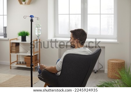 Rare view of male patient receiving intravenous treatment. Young man sitting in armchair during intravenous vitamin therapy in hospital room or wellness center. Medical treatment, healthcare concept Royalty-Free Stock Photo #2245924935