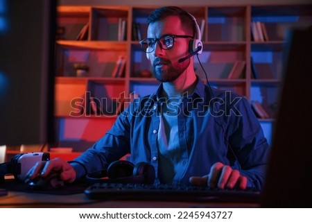 Front view. Man is sitting by a computer indoors at home. Neon lighting.