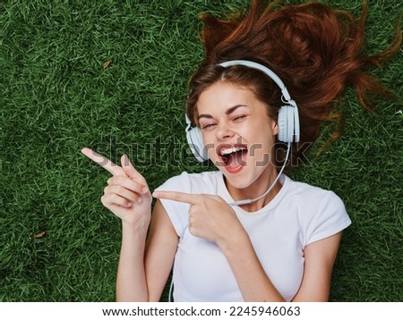 Woman wearing headphones listening to music points the finger to the side, a smile with teeth lies open mouth, lies on the green grass in the summer, good mood happiness Royalty-Free Stock Photo #2245946063