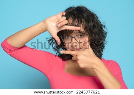 Young brunette woman making a hand frame, composing picture ideas in pink blouse  on a blue studio background. Front view.