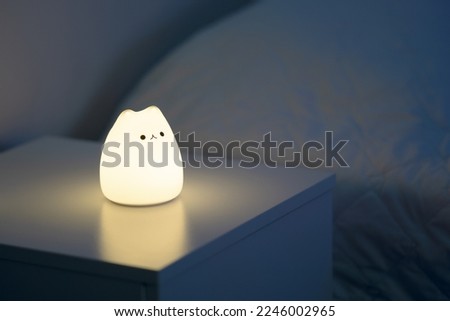 Cute cat shaped night lamp standing on a bedside table next to bed. Bedside lamp. Night lamp standing next to bed. Bedroom lamp on a night table next to a sleeping bed in a dark room. Close up.  Royalty-Free Stock Photo #2246002965
