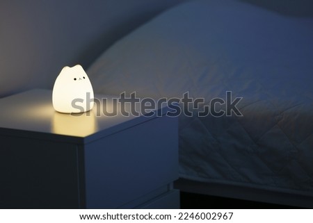 Cute cat shaped night lamp standing on a bedside table next to bed. Bedside lamp. Night lamp standing next to bed. Bedroom lamp on a night table next to a sleeping bed in a dark room.  Royalty-Free Stock Photo #2246002967