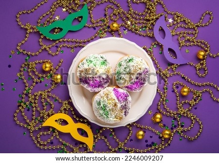 Mardi Gras King Cake sufganiyot donuts, masquerade festival carnival masks, gold beads and golden, green, purple confetti on purple background. Holiday party invitation, greeting card concept.