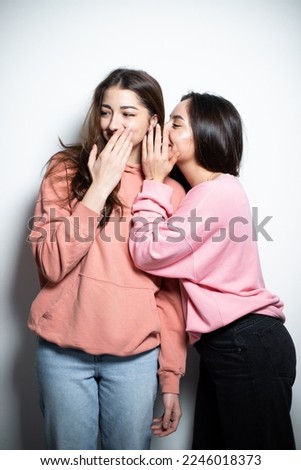 Two young attractive brunette girls in pink sweaters embracing each other, having fun, telling the gossips to each other, smiling, looking at the camera, white background Royalty-Free Stock Photo #2246018373
