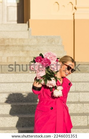 Smiling successful businesswoman in crimson dress and glasses holding bouquet of pink peonies. Small business concept.