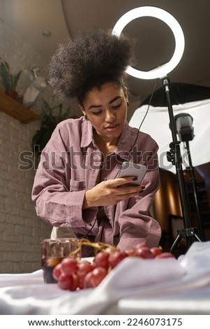 Black female photographer taking photo of cola drink and grapes on white silk cloth on mobile phone at studio. Young girl creating content for photostocks, commerce, social networks and advertising