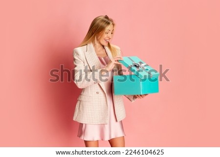 Portrait of beautiful young woman posing, opening present box over pink background. Surprise. Concept of celebration, party, women's day, emotions, holiday, happiness. Ad