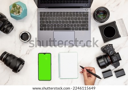 Top view of modern white marble desk of photographer writing in note pad with mobile with green screen chroma key for copy space. Photographer and content creator concept
