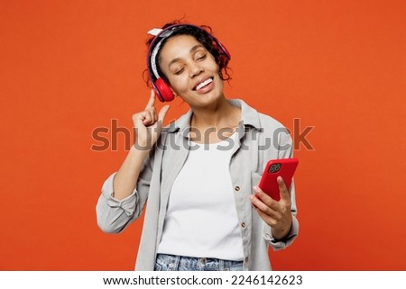 Young woman of African American ethnicity she wears grey shirt headband headphones listen to music use mobile cell phone isolated on plain orange background studio portrait. People lifestyle concept