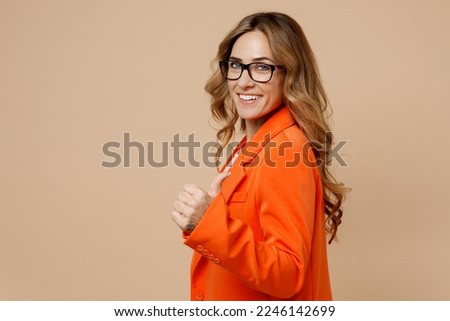 SIde view oung fun successful employee business woman corporate lawyer 30s wear classic formal orange suit glasses work in office showing thumb up like gesture isolated on plain beige color background