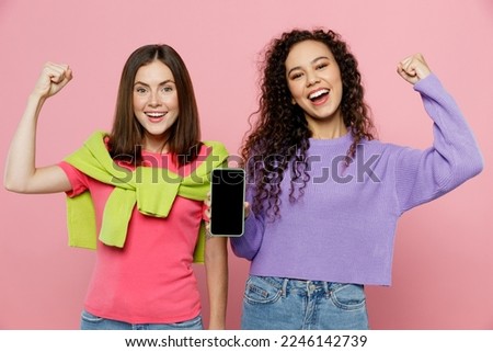 Young two friends women wears green purple shirts together hold in hand use mobile cell phone with blank screen workspace area do winner gesture isolated on pastel plain light pink color background