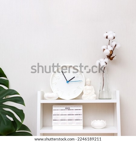Buddha figurine, clock, lotus candlesticks and calendar surrounded by monstera and vases with branch of cotton on white shelf. Concept mental health. Square format, selective focus, copy space