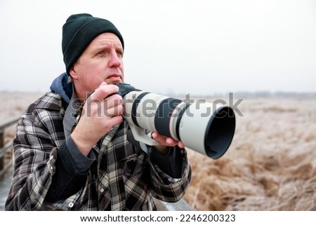 A photographer holds a camera and telephoto lens as the looks out across the winter landscape