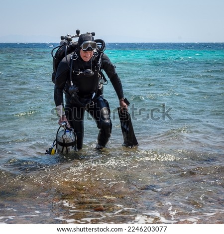 A smiling man diver in scuba gear with one cylinder in his right hand, fins in his left hand and twin cylinders comes out of the sea, after finishing a technical dive Royalty-Free Stock Photo #2246203077
