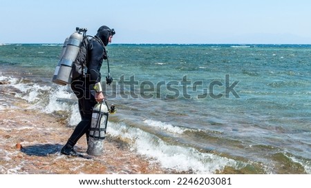 A man diver in scuba gear with one tank in hand and twin tanks enters the sea to start a technical dive Royalty-Free Stock Photo #2246203081