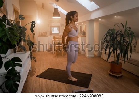 Smiling Asian woman practicing online sports while watching video tutorial on laptop and doing stretching yoga exercises on fitness mat in living room. Training at home. Stretching legs.