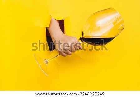 A caucasian man's hand emerges through a torn hole in yellow paper with a large glass of red wine. The concept of alcoholism, drunkenness and hangover. Royalty-Free Stock Photo #2246227249