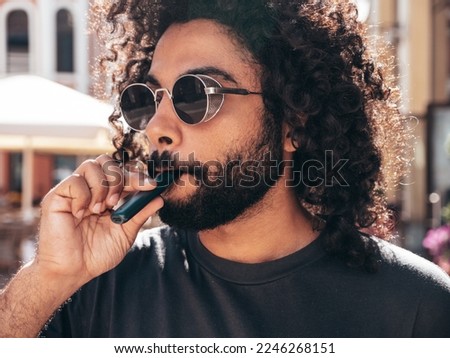 Closeup portrait of handsome smiling hipster model.Unshaven Arabian man smoking electronic cigarette. Fashion male with long curly hairstyle posing in the street. Makes smoke. Steam from vape Royalty-Free Stock Photo #2246268151