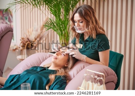 Female eyelashes technician in protective face mask combing woman lashes with disposable mascara brush in beauty salon. Beauty specialist comping client eyelashes after extension procedure. Royalty-Free Stock Photo #2246281169