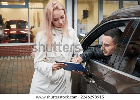 In the yard of car dealership young girl sales consultant carrying out car purchase agreement, signing documents with buyer who sitting in the cabin of his new car.