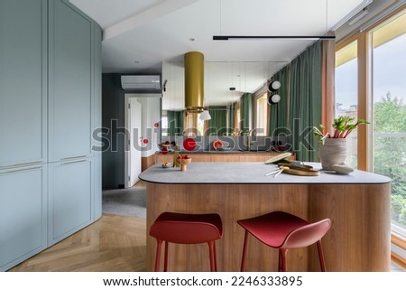 Modern composition of kitchen interior with wooden kitchen island, red barstools, colorful sculpture, green curtain, gold cooker hood, vase with rhubarb and personal accessories. Home decor. Template.