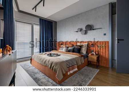 Interior design of elegant bedroom with big orange bed, beige and grey bedclothes, blue curtain, rug, modern lamp, night stand, vase with dried flowers and personal accessories. Home decor. Template. Royalty-Free Stock Photo #2246333901