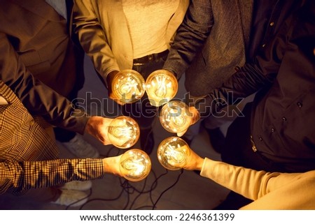 Team of business people share creative energy and innovative ideas. Dark background with cropped shot of human hands holding bright yellow glowing electric Edison lightbulbs. Teamwork, success concept Royalty-Free Stock Photo #2246361399
