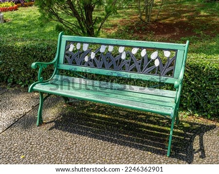 A green park bench in front of a small hedgerow and under a pine tree, with tulip flowers ornament.