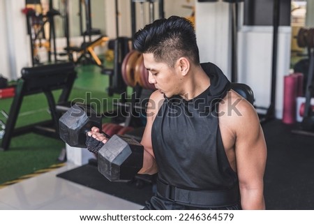 A tanned and focused asian guy doing seated alternate dumbbell curls at the gym. Wearing a hooded low cut tank top. Training biceps at the gym. Royalty-Free Stock Photo #2246364579