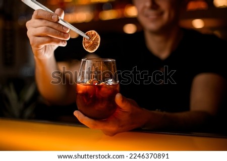hand of male barman neatly holds glass with cold cocktail and garnishes it with slice of dry orange using tweezers Royalty-Free Stock Photo #2246370891
