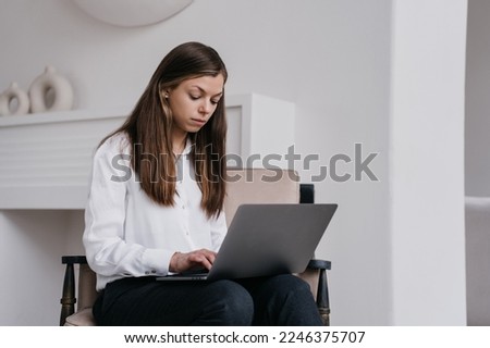 Pensive brunette businesswoman in white shirt and black pants sitting on cozy chair with laptop intense working at office. Serious hispanic female student holds pen, remote learning at home. Student.