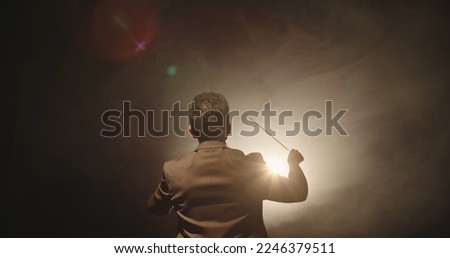 Unrecognizable Male orchestra conductor controlling music in orchestra pit by movement of his hands and white baton, studio shot on black background  Royalty-Free Stock Photo #2246379511