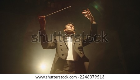 Asian symphony orchestra conductor wearing suit is directing musicians with movement of baton, isolated on black smokey background Royalty-Free Stock Photo #2246379513