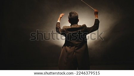 Unrecognizable Male orchestra conductor controlling music in orchestra pit by movement of his hands and white baton, studio shot on black background  Royalty-Free Stock Photo #2246379515