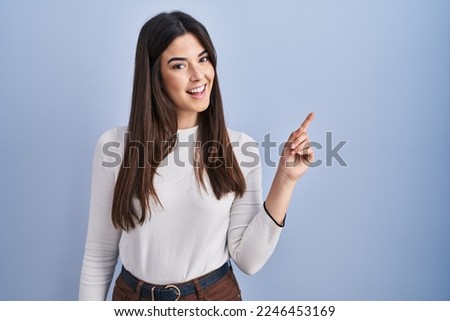 Young brunette woman standing over blue background with a big smile on face, pointing with hand finger to the side looking at the camera.  Royalty-Free Stock Photo #2246453169