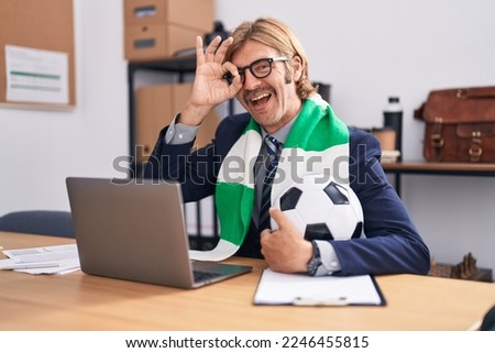 Caucasian man with mustache working at the office supporting football team smiling happy doing ok sign with hand on eye looking through fingers 