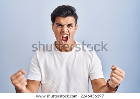 Hispanic man standing over blue background angry and mad raising fists frustrated and furious while shouting with anger. rage and aggressive concept.  Royalty-Free Stock Photo #2246456197