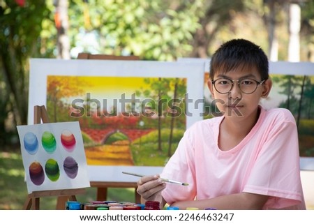 Young Asian teen boy is holding and using paintbrush to draw various pictures on table outside the room happily, raising teen and adult helps kids school project concept.