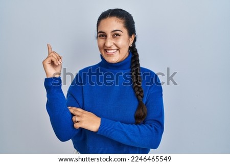 Young hispanic woman standing over isolated background with a big smile on face, pointing with hand finger to the side looking at the camera. 