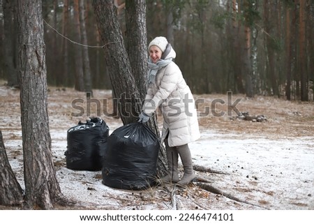 Portrait of happy beautiful elderly senior retired woman in age in forest or park at winter cold day, smiling, picking up garbage bag. Side view.The concept of environment protection and volunteering.