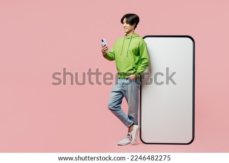 Full body side view young man of Asian ethnicity wear green hoody big huge blank screen mobile cell phone with area hold in hand use smartphone isolated on plain pastel light pink background studio
