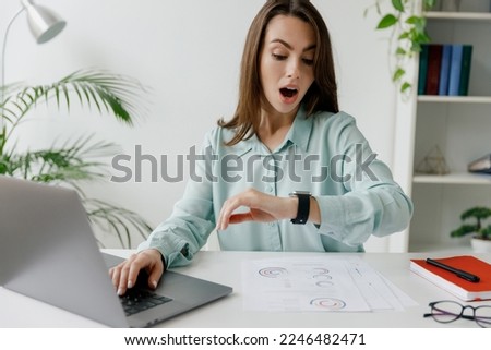 Young confused sad shocked employee business woman 20s wear blue shirt look at smartwatch time sit work at workplace white desk with laptop pc computer at office indoors. Achievement career concept Royalty-Free Stock Photo #2246482471